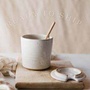 May include: A speckled white ceramic mug with a wooden spoon inside, and a matching lid. The mug is on a wooden cutting board. The text "READY TO SHIP" is arched above the mug.