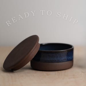 May include: A small, round ceramic container with a dark brown matte base and a glossy, deep blue glazed upper section. The container includes a matching circular, dark brown matte lid that is tilted and resting beside the open vessel.