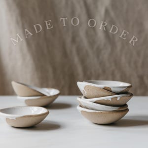 May include: Handmade ceramic bowls with a natural, textured finish and a white glazed rim. The bowls are stacked and arranged on a white surface. The text "MADE TO ORDER" is arched above the bowls.