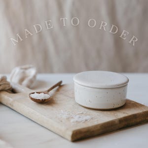 May include: A white ceramic lidded container with brown speckles sits on a wooden cutting board next to a small wooden spoon filled with white salt. The text MADE TO ORDER is visible in the upper portion of the image in a light grey font.