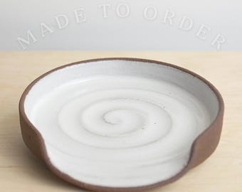 May include: A round ceramic spoon rest with a dark brown exterior and a white interior featuring a spiral pattern. The text MADE TO ORDER is visible in the upper portion of the image. The spoon rest has a curved indentation on one side.