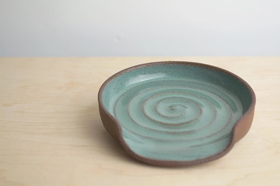 Ceramic spoon rest with glossy green glaze on the inside and dark brown clay showing on the edges and bottom. A swirl design in the clay from the middle to the edges. Piece is about 4.5 inches wide, opening for the spoon in front is about 3 inches.