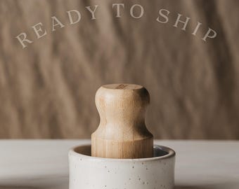 May include: A light-colored wooden pestle sits inside a small, round, white ceramic bowl with dark speckles. The text READY TO SHIP is visible in the upper portion of the image. The pestle has a smooth, contoured handle and a flat base.