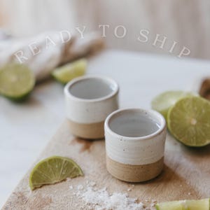 May include: Two small ceramic cups with a white glazed top and a natural tan base, featuring the text READY TO SHIP. The cups are arranged with sliced green limes and coarse salt on a wooden surface, showcasing a simple, cylindrical design.