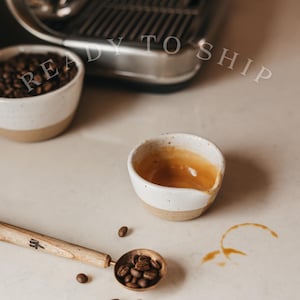 May include: Two small ceramic bowls with a white speckled upper half and a tan lower half, one filled with coffee beans and the other containing espresso. A wooden coffee scoop filled with beans lies nearby. The text READY TO SHIP is visible.