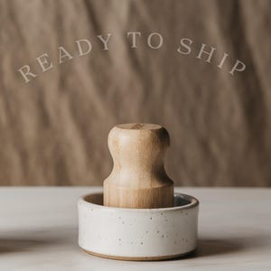 May include: A light-colored wooden muddler with a rounded top is set in a small, speckled white ceramic bowl. The text "READY TO SHIP" is arched above the objects. The background is a neutral brown.