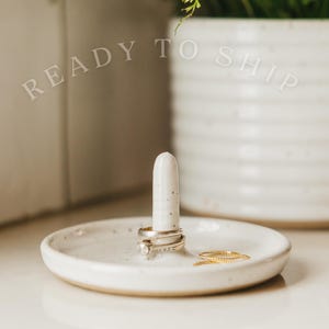 May include: A speckled white ceramic ring holder with a tall, cylindrical post and a shallow dish base. Several rings are displayed on the post and dish. The words "READY TO SHIP" are arched above the ring holder.