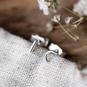 May include: A pair of silver earrings, one is a small hoop and the other is a straight bar, on a white fabric background.
