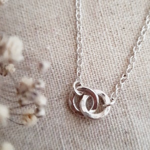 May include: A silver necklace with a pendant of two interlinked rings. The chain is fine, and the rings have a textured surface. The necklace is shown against a neutral fabric background, with soft, blurred white flowers in the corner.