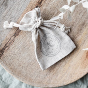May include: A small, white, fabric pouch with a drawstring closure. The pouch has a circular stamp with a design on it.