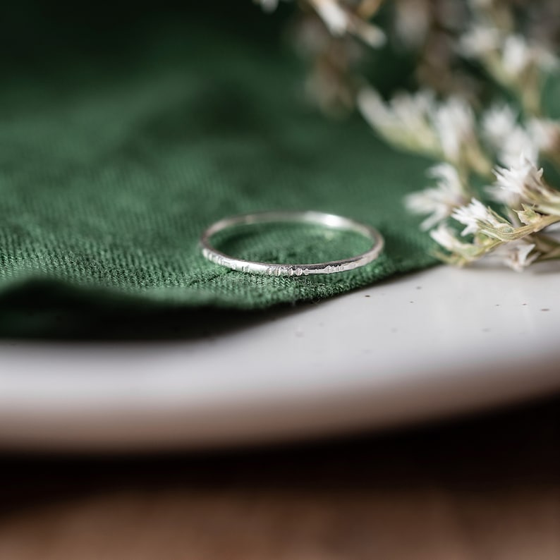 May include: A thin silver ring with a hammered texture, resting on a green fabric surface.