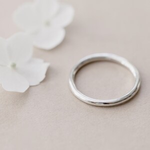 May include: A simple silver band ring with a smooth, rounded surface. The ring is lying on a light brown background.