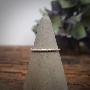 May include: A thin silver band ring with a textured surface. The ring is displayed on a gray concrete ring holder.