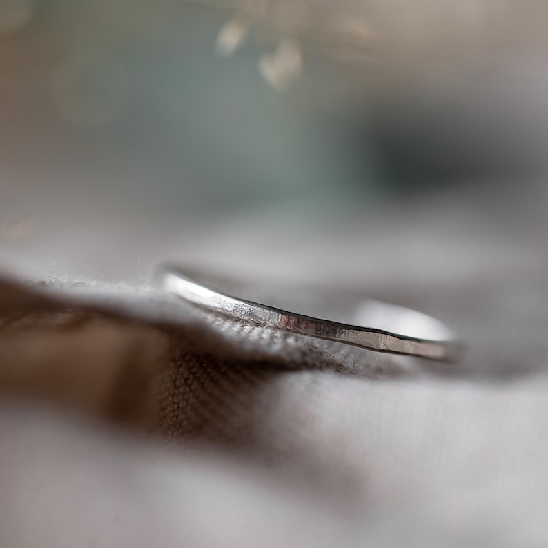May include: A close-up of a silver ring with a textured surface. The ring is thin and has a hammered appearance. The ring is set against a soft, neutral background.