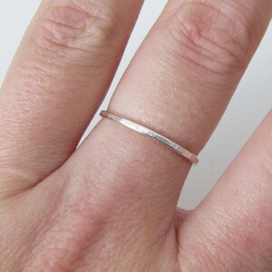 May include: A thin, silver ring with a hammered texture. The ring is shown on a finger, highlighting its delicate design. The band appears to be approximately 1 mm wide, showcasing a minimalist style. The ring's surface reflects light, emphasizing its metallic sheen.