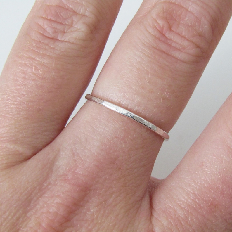 May include: A thin, silver ring with a hammered texture. The ring is shown on a finger, highlighting its delicate design. The band appears to be approximately 1 mm wide, showcasing a minimalist style. The ring's surface reflects light, emphasizing its metallic sheen.