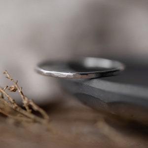 May include: A close-up of a silver ring with a hammered texture. The ring is resting on a dark gray surface, with a blurred background. The ring appears to be made of metal.