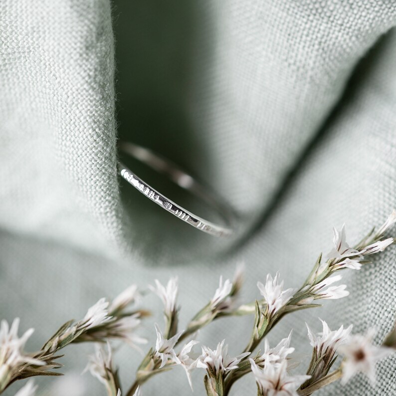 May include: A thin silver band ring with a textured surface, resting on a light green fabric background with white flowers.