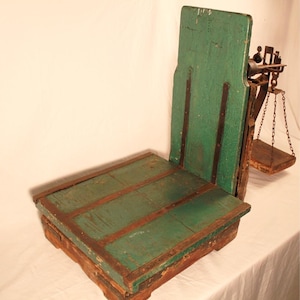 May include: An antique green and brown wooden scale with a platform base and a vertical back. The scale has a metal balance arm with a small tray suspended by chains. The wood shows signs of wear.