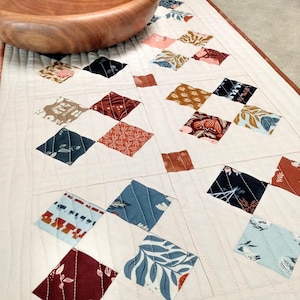 Pode incluir: Um corredor de mesa branco com uma borda marrom, apresentando um padrão de diamante de quadrados de tecido coloridos com desenhos florais e geométricos.