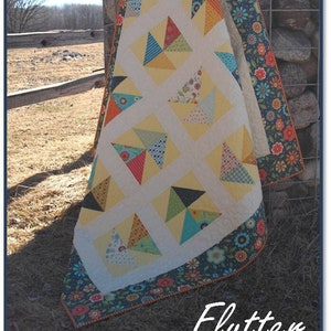 May include: A quilt with a white background and a patchwork design of colourful triangles. The quilt is bordered with a dark blue fabric with a floral pattern. The quilt is draped over a stone wall and the text "Flutter" is written in white below the quilt.
