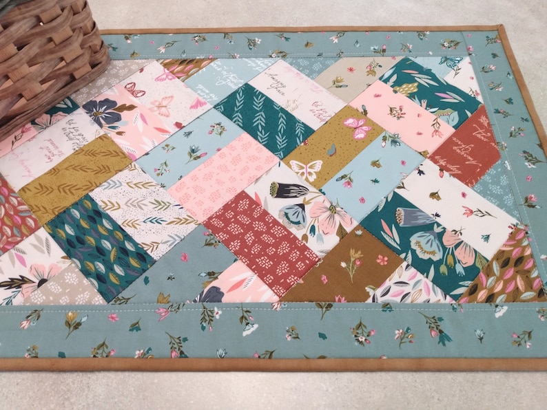 May include: A quilted table runner with a diamond pattern featuring various floral and leaf prints in shades of pink, green, brown, and blue. The runner has a light blue border with a floral pattern and a brown suede backing.