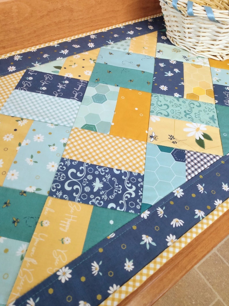 May include: A patchwork quilt table runner with a bee and flower theme. The runner is made of various fabrics in shades of blue, yellow, and green. The runner has a yellow and white gingham border and a blue border with white daisies. The runner features the text "Be Happy, Be You, Be Strong".