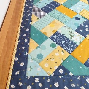 May include: A blue and yellow table runner with a patchwork design featuring bees, flowers, and geometric patterns. The runner has a white daisy print border.