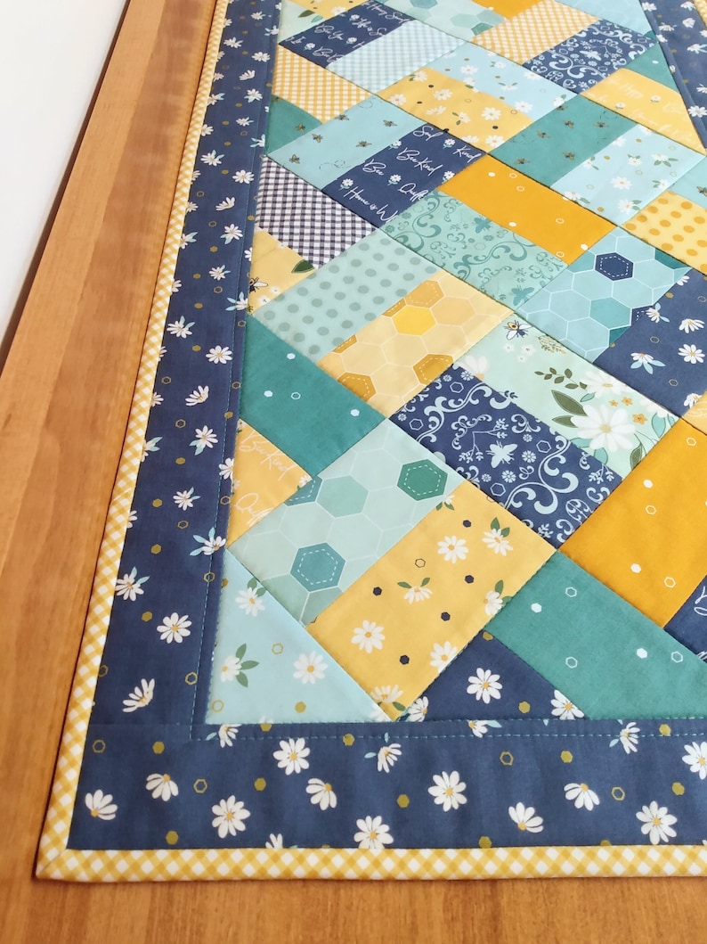 May include: A blue and yellow table runner with a patchwork design featuring bees, flowers, and geometric patterns. The runner has a white daisy print border.
