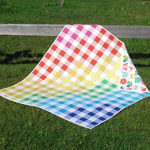 May include: A colorful picnic blanket with a rainbow gingham pattern. The blanket transitions from pink to red, yellow, green, and blue. The reverse side features a pattern of cartoon characters. The blanket is spread out on green grass.