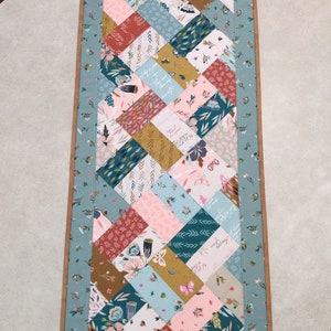 May include: A teal and brown table runner with a patchwork design featuring floral and leaf patterns. The runner is made of fabric squares sewn together in a diamond pattern. The fabric squares feature various colors, including teal, pink, brown, and gold. The runner has a teal border with a brown outer border.