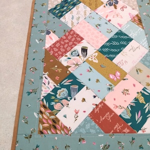 May include: A teal blue table runner with a brown border. The runner is made of fabric with a patchwork design featuring various floral patterns, butterflies, and text. The text includes "Amazing Grace" and "Your Love".