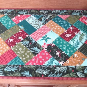 May include: A festive table runner with a patchwork design featuring a variety of holiday-themed fabrics. The runner is made up of squares and rectangles in shades of red, green, blue, and gold. The fabric patterns include snowflakes, pine branches, and birds.