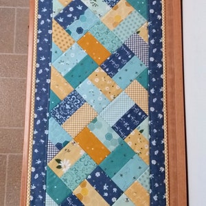 May include: A blue and yellow patchwork table runner with a white floral border. The runner is made of various fabrics with different patterns, including polka dots, gingham, and floral prints.