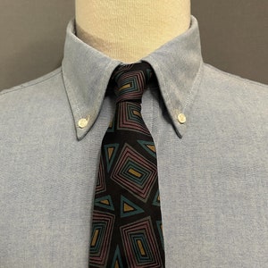 Puede incluir: Una corbata negra con un patrón geométrico de cuadrados y triángulos en tonos de azul verdoso, rosa y dorado. La corbata se lleva con una camisa de botones azul claro.