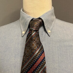 May include: A light blue button-down shirt with a patterned tie. The tie has a dark blue, red, and gold pattern.