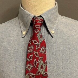 May include: A red tie with a repeating diamond pattern in white, black, and gray is knotted against a light blue button-down shirt. The tie's design is a geometric pattern.