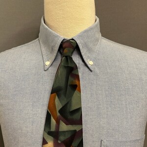 Puede incluir: Una camisa azul claro con botones, bolsillo y corbata estampada. La corbata presenta un diseño geométrico en tonos verdes, marrones y morados. La camisa se exhibe en un maniquí.