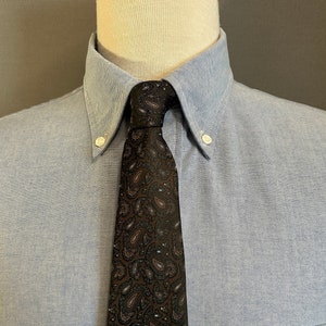 May include: A light blue button-down shirt with a black paisley patterned tie. The shirt has a pocket on the left side.