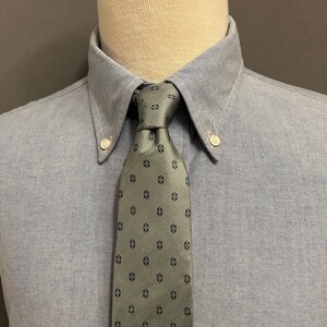 Könnte beinhalten: Ein hellblaues Button-Down-Hemd mit einer grauen Krawatte. Die Krawatte hat ein sich wiederholendes Muster aus kleinen, dunkelblauen geometrischen Formen. Der Hemdkragen hat Button-Down-Spitzen und weiße Knöpfe. Das Hemd befindet sich auf einer Schaufensterpuppe.