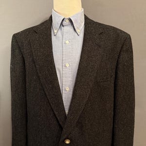 May include: A dark gray herringbone blazer with a light blue button-down shirt. The blazer has a two-button closure and a notched lapel. The shirt has a button-down collar. The outfit is displayed on a mannequin.