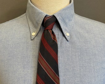 Peut inclure: Une chemise boutonnée bleu clair avec une cravate rayée. La cravate présente des rayures alternées rouges, marron et beige sur un fond sombre. La chemise a un col boutonné et une poche poitrine.
