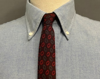 Peut inclure: Gros plan d'une chemise bleu clair boutonnée avec une cravate à motifs. La cravate présente un motif répétitif d'ovales rouges et gris sur un fond bordeaux foncé. Le col de la chemise est boutonné.