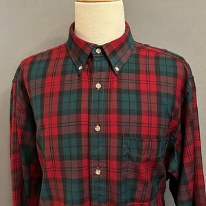 以下が含まれることがあります： 赤と緑の格子柄の長袖ボタンダウンシャツ。シャツはボタンダウンカラーで、左側にポケットが付いています。