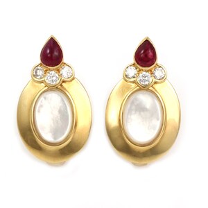May include: A pair of gold earrings with oval-shaped mother-of-pearl stones set in a gold frame. Each earring features a ruby and three diamonds.
