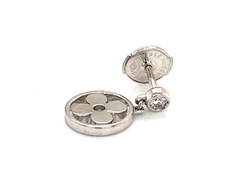 K&ouml;nnte beinhalten: Ein silberner Ohrring mit einem baumelnden Diamanten und einem runden Anh&auml;nger mit einem ausgeschnittenen vierbl&auml;ttrigen Blumendesign. Die R&uuml;ckseite des Ohrrings tr&auml;gt die Aufschrift "Louis Vuitton Paris".