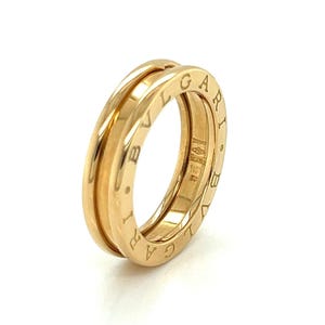 May include: A gold-colored ring with a double-band design. The ring has engraved lettering around the outer band. The ring is made of a shiny, reflective metal.