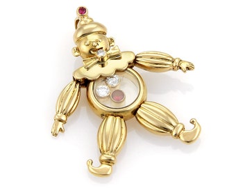 May include: A gold-toned clown pendant with a ruby nose and diamond accents. The clown's face is visible through a clear circular window, revealing a small red ruby and diamonds.