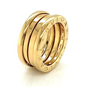 May include: A gold-colored ring with a multi-band design. The ring features three bands that are stacked together. The outer band has engraved lettering. The ring is set against a white background.