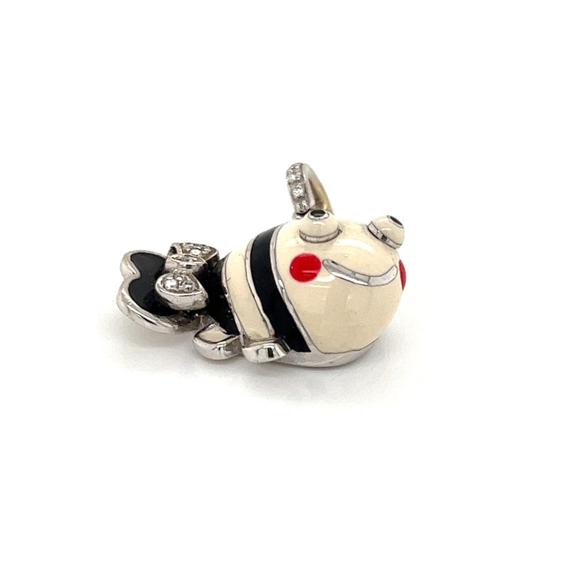 May include: A white and black enamel charm with red accents, shaped like a cartoon fish with a diamond-encrusted bow. The charm is made of silver and has a loop for attaching to a necklace or bracelet.